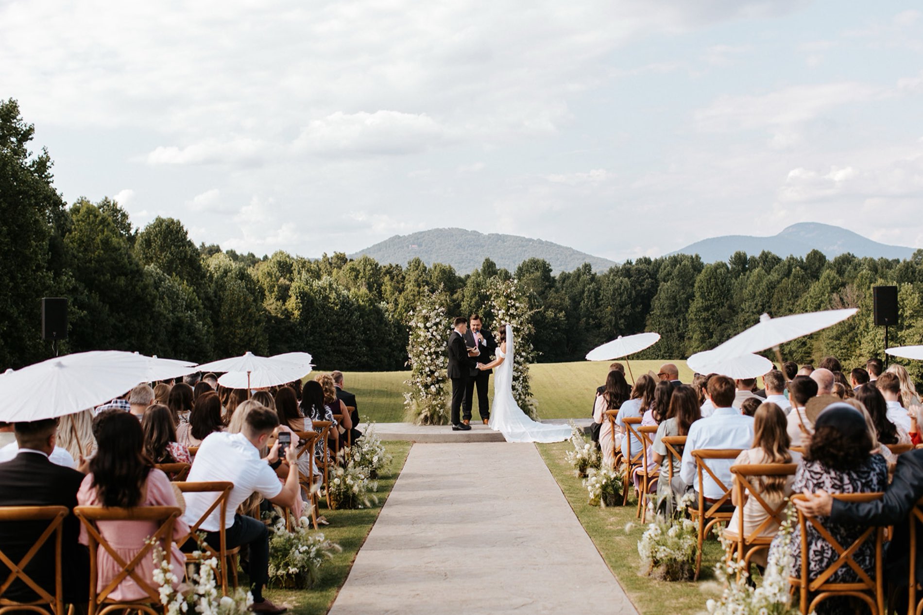 A ceremony seen from behind the guests, white parasols raised, mountains beyond the arch